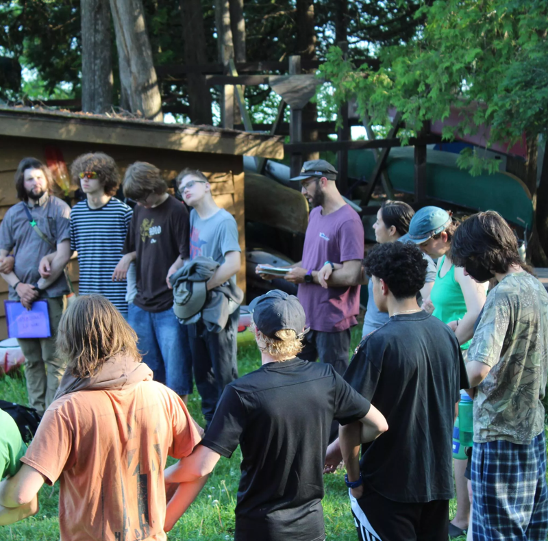 A group of about a dozen young people are gathered outdoors, seemingly listening to a man who is holding a book or papers. They are standing in a semi-circle on a grassy area, with some kind of structure or shed visible in the background. The atmosphere appears casual and informal, possibly a class or a meeting in a relaxed setting.